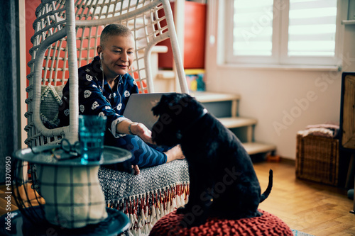 Smiling cute senior woman with short hair sitting in her chair with laptop in her lap and playing with her dog.