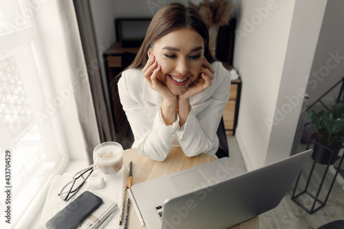 Beautiful young businesswoman, woman using laptop and smiling while working indoors. Home office during Coronavirus or Covid-19 quarantine. Communicates on internet.