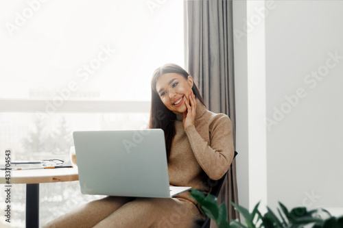 Beautiful young woman in casual clothing using laptop and smiling while working indoors. Home office during Coronavirus or Covid-19 quarantine. Communicates on internet.