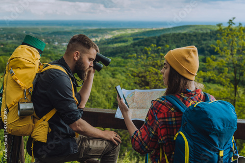 Young couple of hikers using a smartphone and a map while standing on a lookout with camping gear
