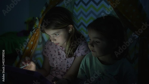 Girls look at the tablet at home at night. Sisters. sitting in a wigmam in the home room and watching sultics on the tablet. Children's lives at home. photo