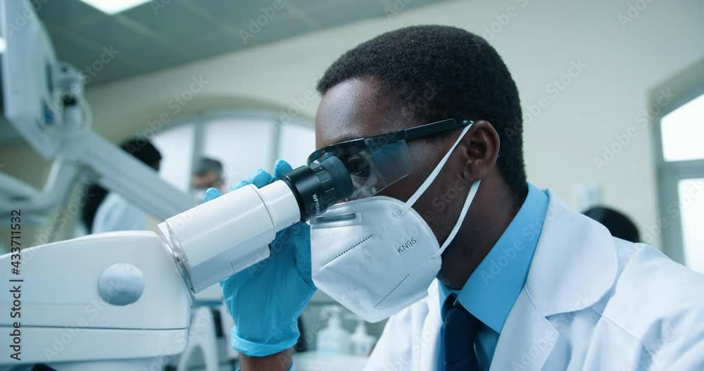 Close up of African American young man scientist working in labs on cure against Covid spreading. Chemical expert in mask looking at analysis through microscope, developing new medication, experiment Close up of African American young man scientist working in labs on cure against Covid spreading. Chemical expert in mask looking at analysis through microscope, developing new medication, experiment