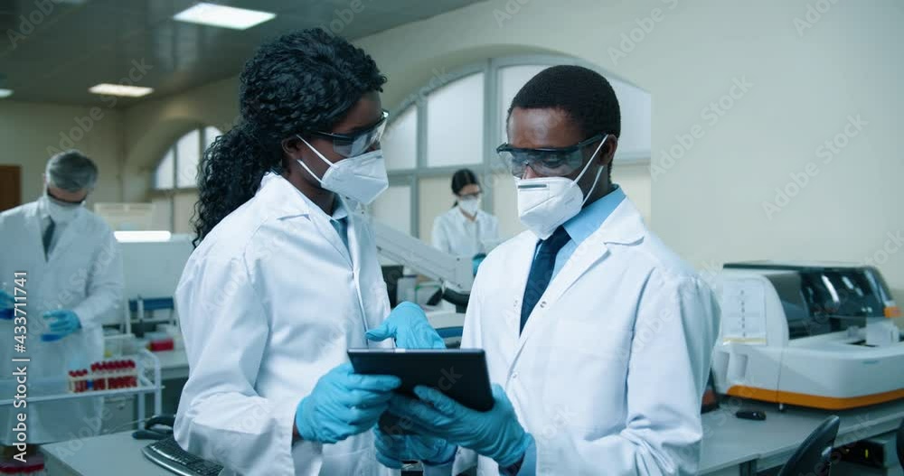 Portrait of African American young male and female medical experts in masks and white coats standing in clinic laboratory and tapping on tablet discussing analysis results. Healthcare, lab concept Portrait of African American young male and female medical experts in masks and white coats standing in clinic laboratory and tapping on tablet discussing analysis results. Healthcare, lab concept