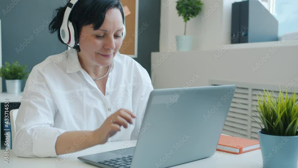 Joyful mature businesswoman is working with laptop typing and enjoying music through headphones in office. Entertainment and workplace concept. Joyful mature businesswoman is working with laptop typing and enjoying music through headphones in office. Entertainment and workplace concept.