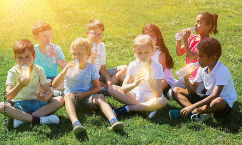 Group of happy kids drinking water and sitting on grass in park outdoors