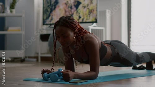 Athlete african in sportwear doing fitness exercices putting cronometer on phone sitting on yoga map in living room. Flexible woman stretching muscle body enjoying healthy lifestyle photo