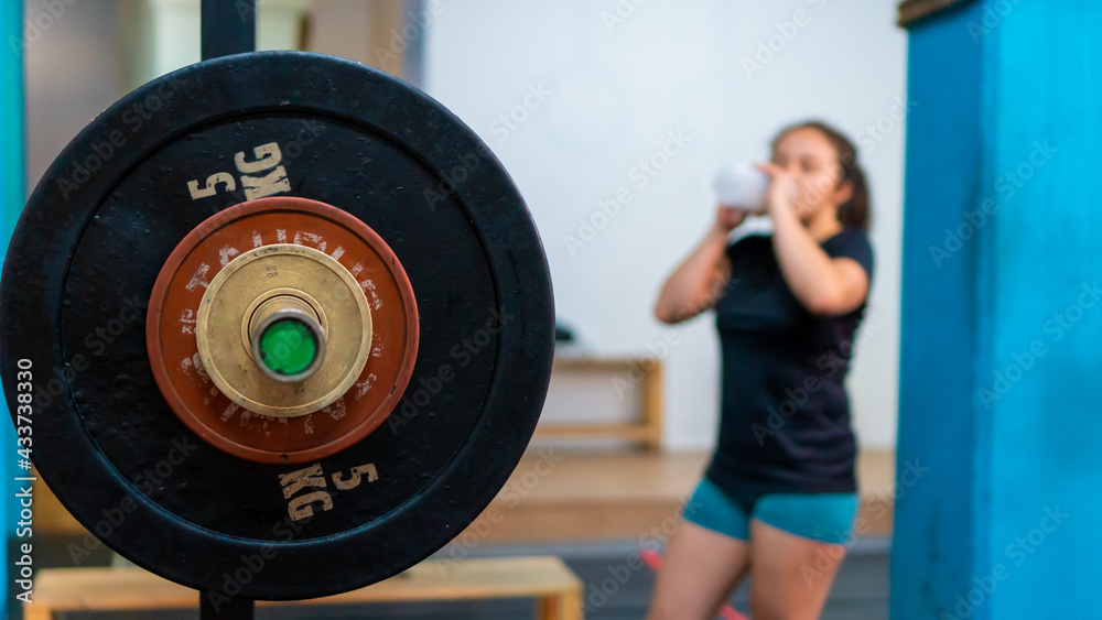 detail photo of a heavy weight on a barbell, with an out-of-focus woman drinking water in the background in a gymnasium.
