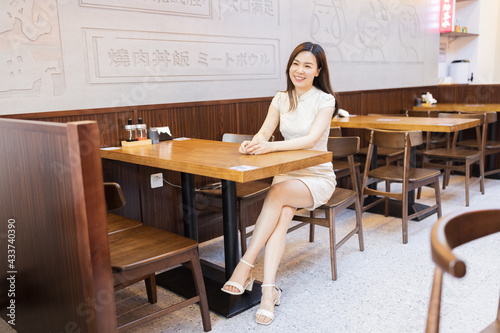 Beautiful young asian woman in traditional white dress named cheongsam sitting in Japanese restaurant or cafeteria and waiting ordered food. Restaurant interior decorated with colorful paper lanterns