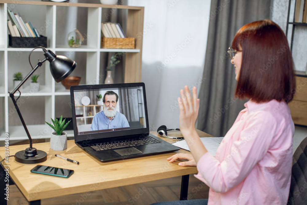 Smiling young woman talking and waving with hand during video call on laptop. Pretty granddaughter chatting online with senior grandfather while staying at home during pandemic time. Smiling young woman talking and waving with hand during video call on laptop. Pretty granddaughter chatting online with senior grandfather while staying at home during pandemic time.