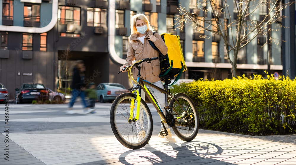 Portrait of beautiful woman delivery worker in medical mask for riding bike standing at street. Pretty female courier with backpack. Food delivery concept