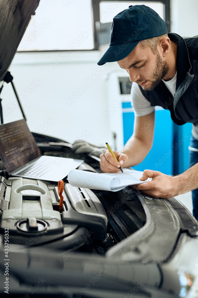 Auto mechanic writing notes while running car diagnostic and examining engine at repair shop. Auto mechanic writing notes while running car diagnostic and examining engine at repair shop.