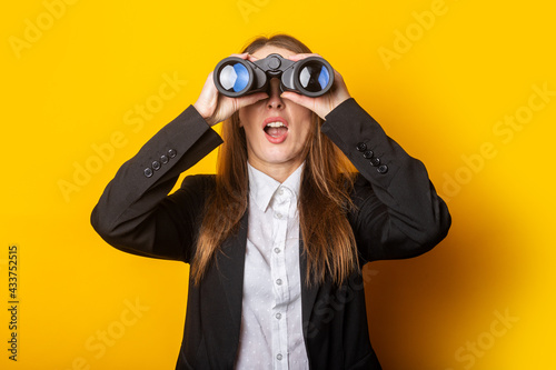 surprised young business woman looking through binoculars on yellow background