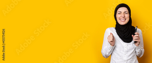 cheerful joyful young woman in hijab holds a phone on a yellow background. Banner © Alex