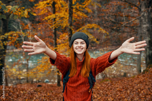 happy woman with a backpack travels in the park in autumn and gesticulate with the hands of the river in the background