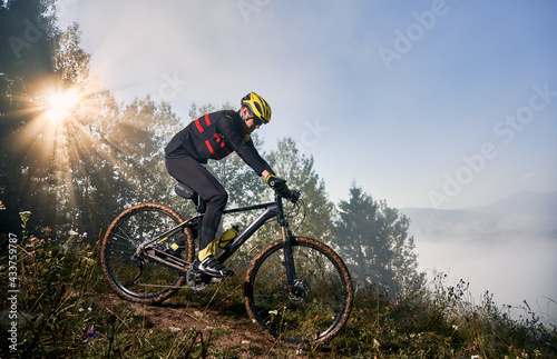 Fototapeta Naklejka Na Ścianę i Meble -  Horizontal snapshot of man riding his bike in the mountains in early foggy morning going downhill. Sun is shining through tree crown, peaks in clouds on background. Concept of active lifestyle