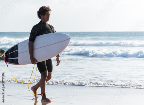 Young malagasy african surfer walking by the beach shore holding his surfboard