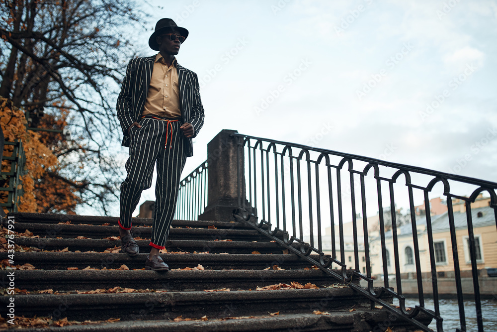 Elegant young African American man wearing stylish striped suit, black hat and glasses going down concrete steps walking by city river, keeping his hands in pockets man walk city