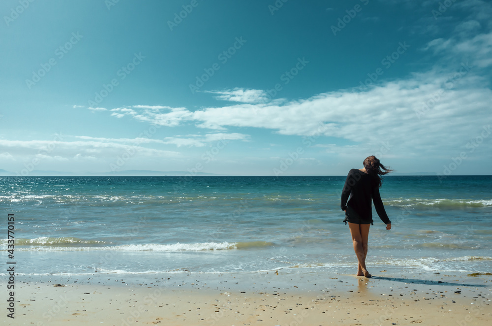 Woman enjoying the warm breeze at the seaside wading in the surf at the edge of a sandy tropical beach with the breeze blowing her hair Woman enjoying the warm breeze at the seaside wading in the surf at the edge of a sandy tropical beach with the breeze blowing her hair