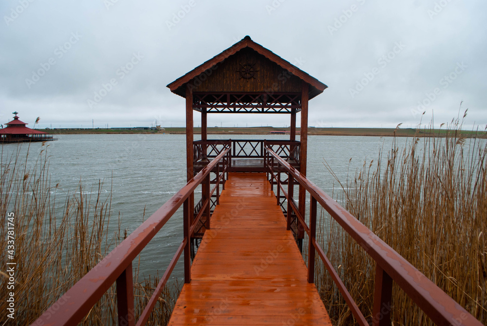 The image of a dark red cabin on the lake surrounded by reeds. The red bridge leading to a cabin on the lake. Lake, reeds, small red cabin