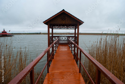 The image of a dark red cabin on the lake surrounded by reeds. The red bridge leading to a cabin on the lake. Lake  reeds  small red cabin