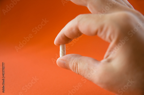 Hand and fingers holding medicine capsule in front of orange background. selective focus pill.