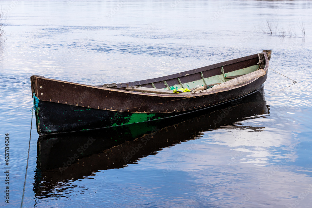 Small brown green wooden fishing row boat at coast. Weathered wooden boat reflecting in blue calm water. Fishing vessel ready to go. Small brown green wooden fishing row boat at coast. Weathered wooden boat reflecting in blue calm water. Fishing vessel ready to go.