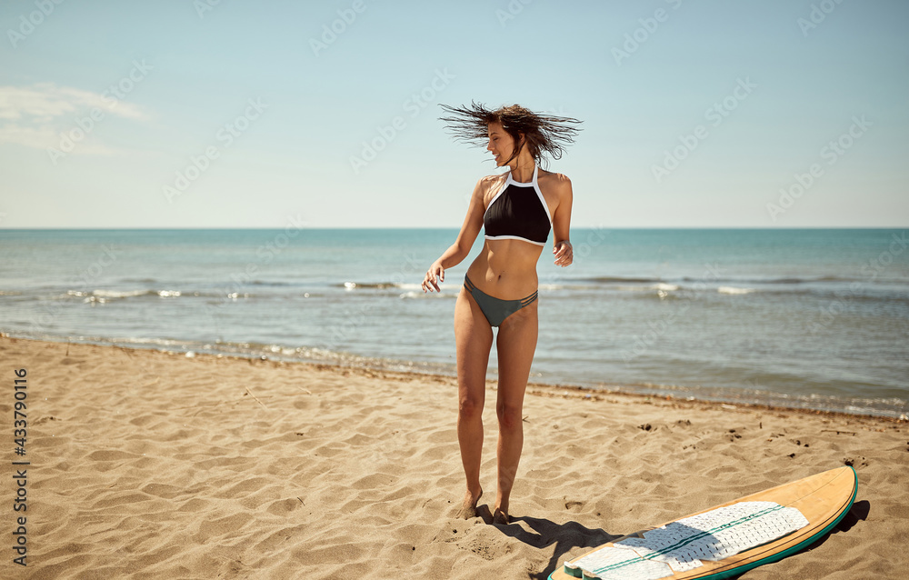 Naklejka premium Young sexy girl with a surfboard on the beach shakes water from her hair while enjoying at sea. Summer, vacation, sea