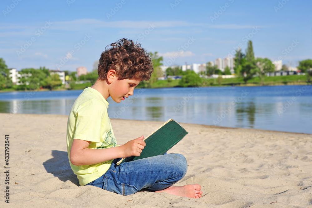 Cute little boy reads an old book. He sits on the sand at the beach