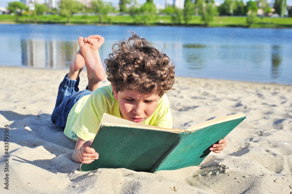 Cute little boy reads an old book. He lies on the sand on the beach