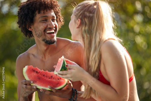  multiethnic couple looking each other  smiling  laughing  while eating watermelon