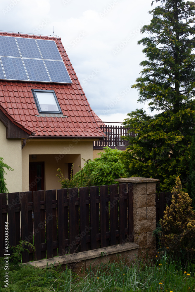 Le pignon d´une maison, la toiture est recouverte de panneaux solaires, il y a une fenêtre velux sur le toit. Au premier plan se trouve une palissade et une haie végétale.