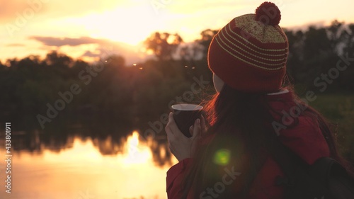 Woman Traveler drinks hot coffee from a mug and looks at the sunset by the river. Free girl traveling admiring the landscape. A tourist is drinking tea from a mug in the sun. travel, hiking