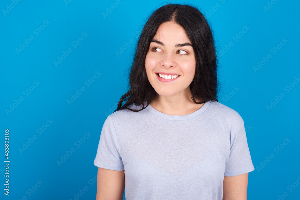 Amazed young beautiful tattooed girl wearing blue t-shirt standing against blue background bitting lip and looking tricky to empty space. Amazed young beautiful tattooed girl wearing blue t-shirt standing against blue background bitting lip and looking tricky to empty space.