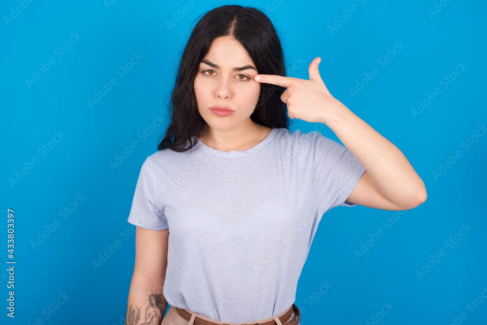 Unhappy young beautiful tattooed girl wearing blue t-shirt makes suicide gesture and imitates gun with hand, curves lips keeps two fingers on temple, shoots, being tired of everything, Unhappy young beautiful tattooed girl wearing blue t-shirt makes suicide gesture and imitates gun with hand, curves lips keeps two fingers on temple, shoots, being tired of everything,