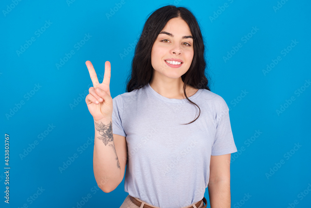 young beautiful tattooed girl wearing blue t-shirt standing against blue background showing and pointing up with fingers number two while smiling confident and happy. young beautiful tattooed girl wearing blue t-shirt standing against blue background showing and pointing up with fingers number two while smiling confident and happy.