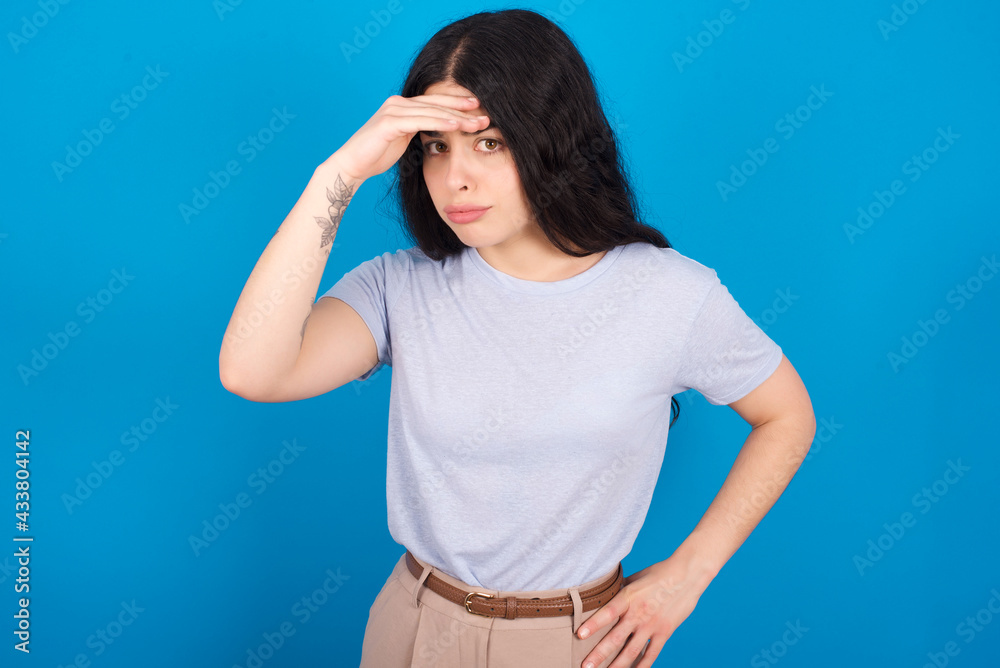 young beautiful tattooed girl wearing blue t-shirt standing against blue background having problems, worried and stressed holds hand on forehead. young beautiful tattooed girl wearing blue t-shirt standing against blue background having problems, worried and stressed holds hand on forehead.