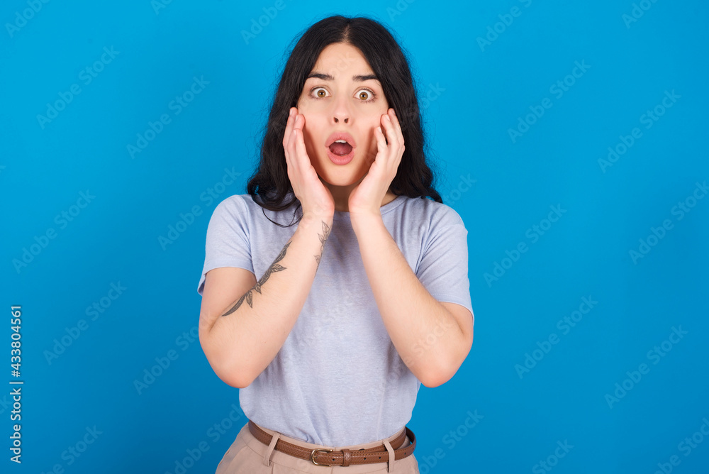 Scared terrified young beautiful tattooed girl wearing blue t-shirt standing against blue background shocked with prices at shop, People and human emotions concept Scared terrified young beautiful tattooed girl wearing blue t-shirt standing against blue background shocked with prices at shop, People and human emotions concept