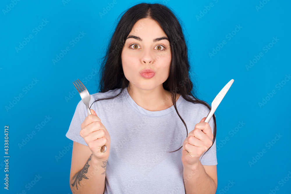 hungry young beautiful tattooed girl wearing blue t-shirt standing against blue background holding in hand fork knife want tasty yummy pizza pie hungry young beautiful tattooed girl wearing blue t-shirt standing against blue background holding in hand fork knife want tasty yummy pizza pie
