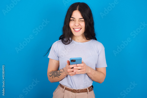 young beautiful tattooed girl wearing blue t-shirt standing against blue background taking a selfie  celebrating success