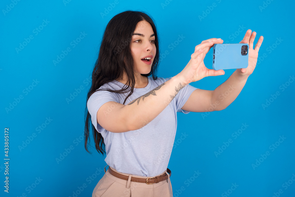 Fototapeta premium young beautiful tattooed girl wearing blue t-shirt standing against blue background taking a selfie to post it on social media or having a video call with friends.