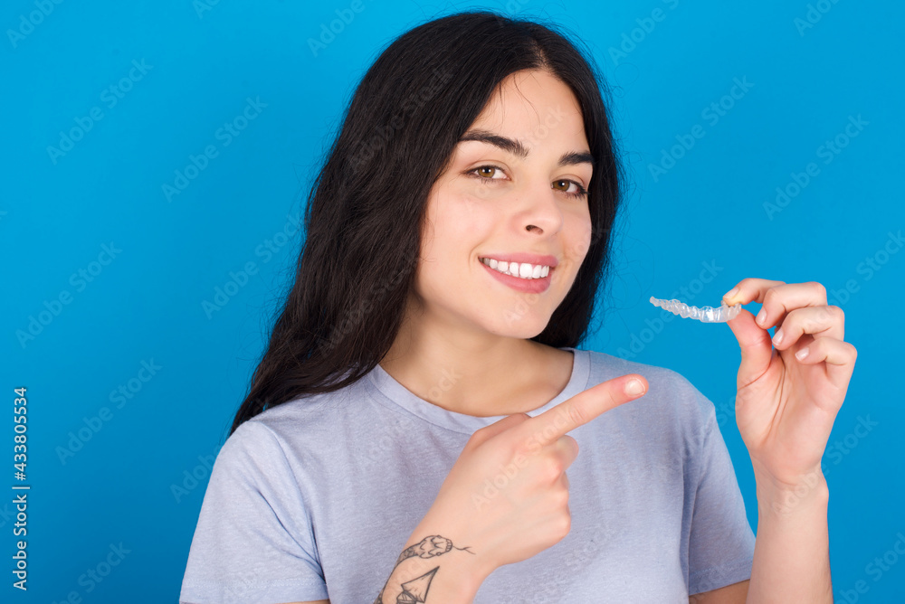 young beautiful tattooed girl wearing blue t-shirt standing against blue background holding an invisible aligner and pointing at it. Dental healthcare and confidence concept. young beautiful tattooed girl wearing blue t-shirt standing against blue background holding an invisible aligner and pointing at it. Dental healthcare and confidence concept.