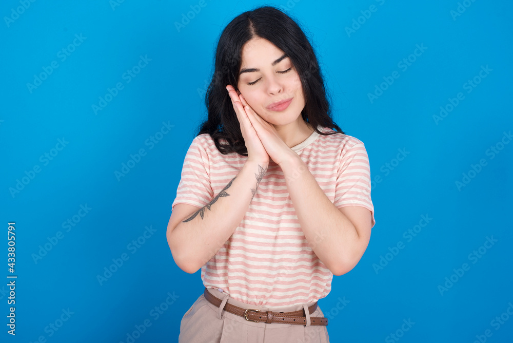 young beautiful tattooed girl wearing pink striped t-shirt standing against blue background leans on pressed palms closes eyes and has pleasant smile dreams about something young beautiful tattooed girl wearing pink striped t-shirt standing against blue background leans on pressed palms closes eyes and has pleasant smile dreams about something