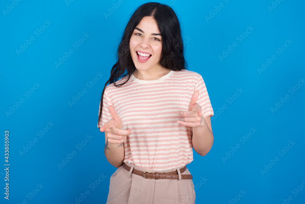 young beautiful tattooed girl wearing pink striped t-shirt standing against blue background directs fingers at camera selects someone. I recommend you. Best choice young beautiful tattooed girl wearing pink striped t-shirt standing against blue background directs fingers at camera selects someone. I recommend you. Best choice