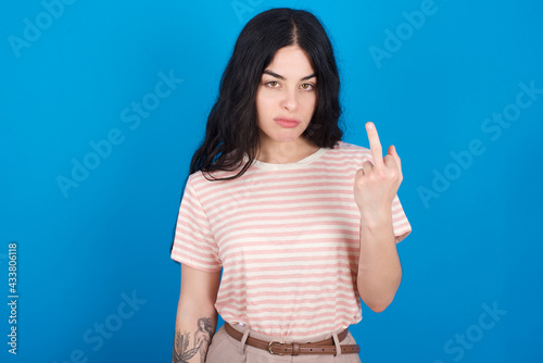 young beautiful tattooed girl wearing pink striped t-shirt standing against blue background shows middle finger bad sign asks not to bother. Provocation and rude attitude.