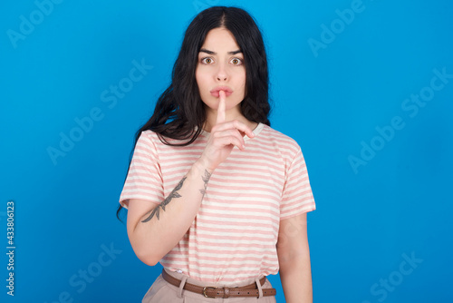 Surprised young beautiful tattooed girl wearing pink striped t-shirt standing against blue background makes silence gesture, keeps finger over lips and looks mysterious at camera