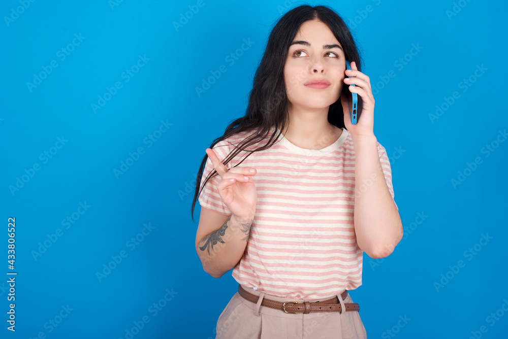 young beautiful tattooed girl wearing pink striped t-shirt standing against blue background speaks on mobile phone spends free time indoors calls to friend. young beautiful tattooed girl wearing pink striped t-shirt standing against blue background speaks on mobile phone spends free time indoors calls to friend.