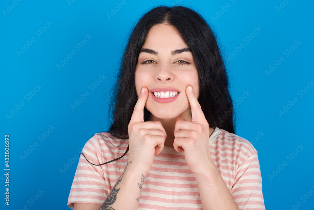 Happy young beautiful tattooed girl wearing pink striped t-shirt standing against blue background with toothy smile, keeps index fingers near mouth, fingers pointing and forcing cheerful smile