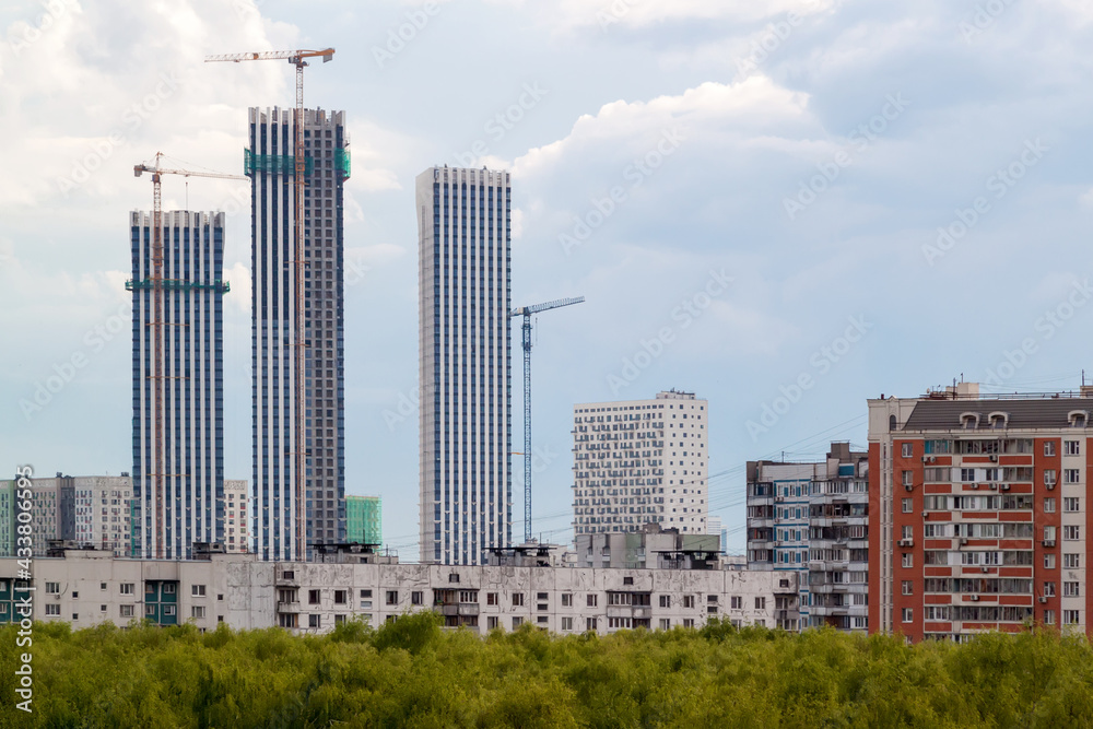 View of High construction buildings site, industrial cranes and apartment buildings at green area. Construction in the city in the green zone with white clouds in blue sky.