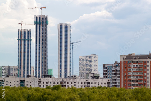 View of High construction buildings site, industrial cranes and apartment buildings at green area. Construction in the city in the green zone with white clouds in blue sky.