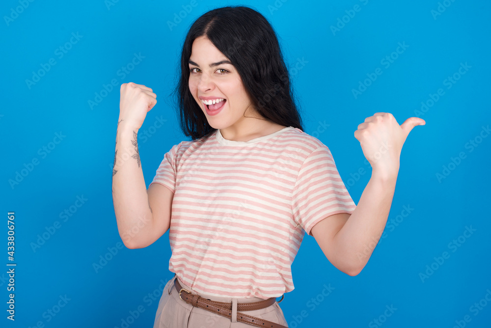 Hooray cool young beautiful tattooed girl wearing pink striped t-shirt standing against blue background point back empty space hand fist Hooray cool young beautiful tattooed girl wearing pink striped t-shirt standing against blue background point back empty space hand fist